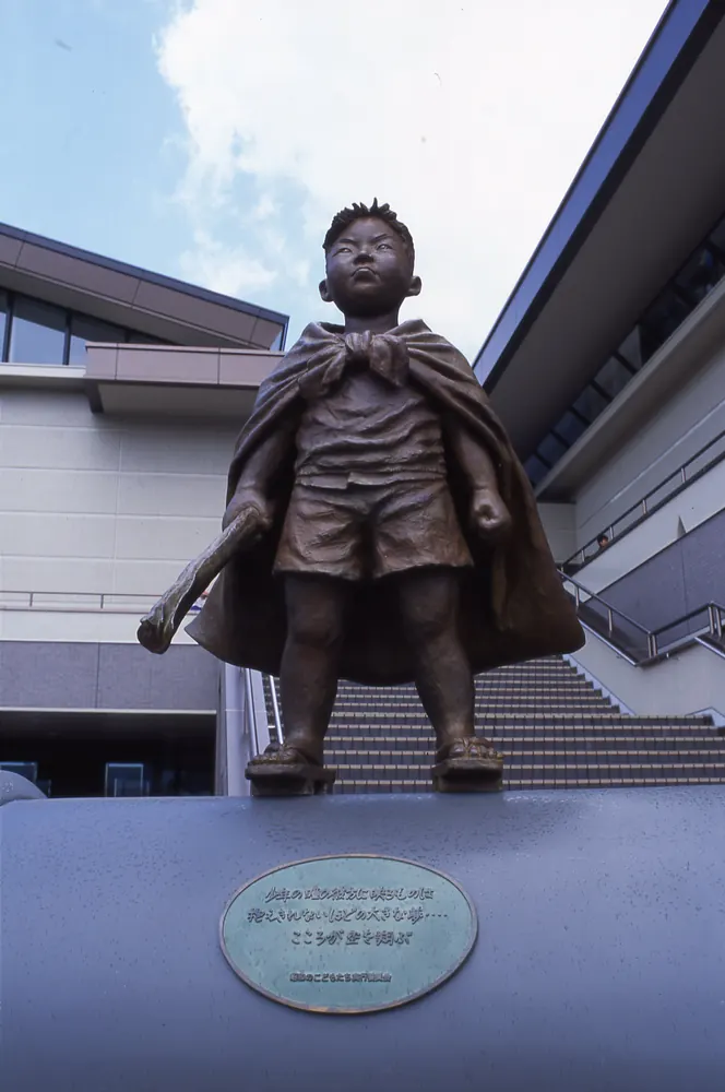 建物の前の台の上に立つ、マントをはおった男の子のブロンズオブジェ「わんぱく」